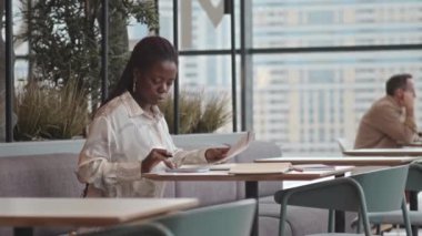 Kafedeki panoramik pencereli masada oturan genç Afrikalı Amerikalı kadın iş belgelerini analiz ediyor.