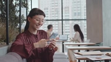 Kafedeki kafeteryada panoramik pencereli masada oturmuş akıllı telefondan mesaj atarken gülümseyen beyaz iş kadını.