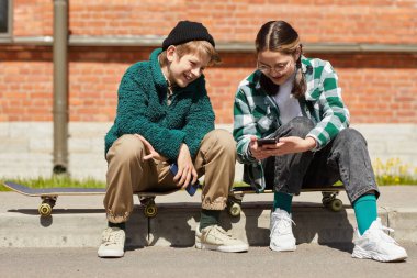 İki arkadaş kaykaylarının üzerinde oturup, dışarıda gezdikten sonra cep telefonundan komik videolar izliyorlar.