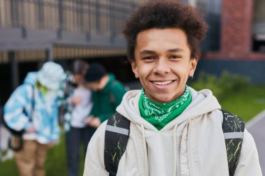 Portrait of African teenage boy with backpack behind his back smiling at camera standing outside of school building clipart