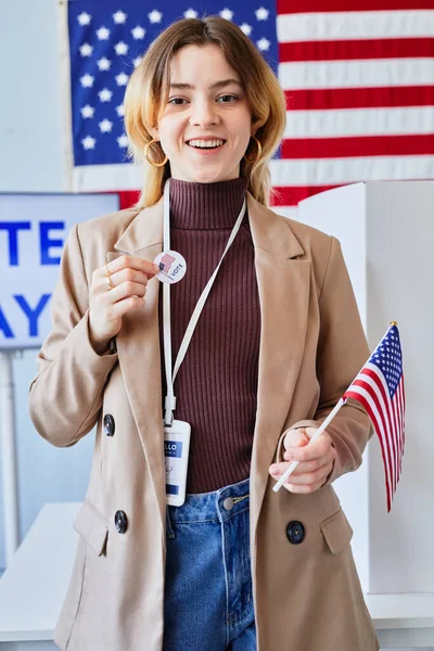 Vertical portrait of smiling young woman holding I vote sticker while standing in voting station with American flag