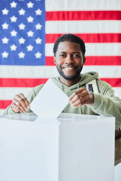 バックグラウンドでアメリカの旗に対してビンに投票する笑顔の黒人男性の垂直な肖像画 — ストック写真