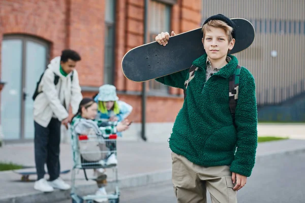スケートボードを保持しているスケーターボーイの肖像画とバックグラウンドで友人とストリートに立っている間のカメラを見る — ストック写真