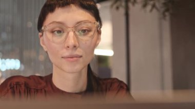 İçerideki kafede dizüstü bilgisayarla çalışan gözlüklü, konsantre olmuş, beyaz bir kadın.