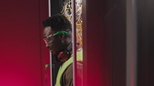 データセンターのラックサーバーキャビネットに対して立っているカメラで笑顔で安全眼鏡とヘッドフォンで黒人の男性Itエンジニアの肖像画をウエスト — ストック動画
