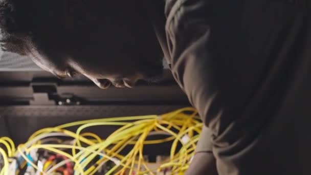 データセンターのダークサーバールームでワイヤーを接続する若い黒人男性It技術者の垂直ショット — ストック動画