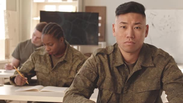 軍事アカデミーで勉強している教室の机に座っている間 カメラを見ているカモフラージュのユニフォームの若いアジアの男性キャデットのスローモの肖像画を待つ — ストック動画
