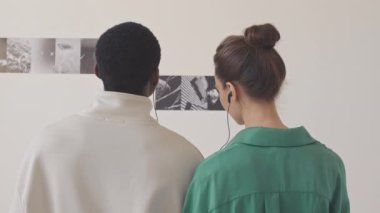 Modern sanat galerisindeki duvarlardaki soyut siyah ve beyaz resimlere bakarken kulaklıkla müzik rehberi dinleyen genç ve etnik çeşitlilikte bir çiftin arka plan görüntüsü