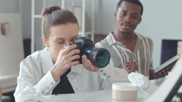 集中的年轻白人女性摄影师使用数码相机拍摄白色背景的护肤产品 与非裔美国男性助手在明亮的照相馆工作 — 图库视频影像