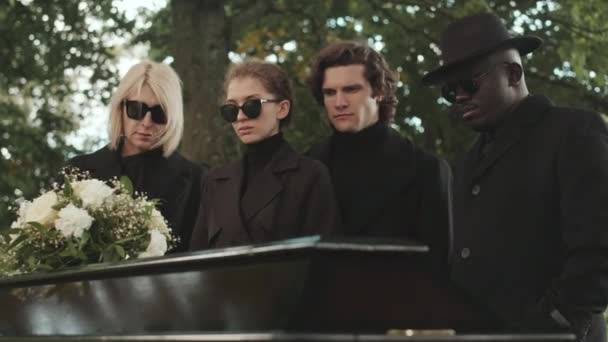 屋外の葬儀で白い花の花束で黒い棺を見ながら悲しんでいる4人の多民族の中型ショット — ストック動画