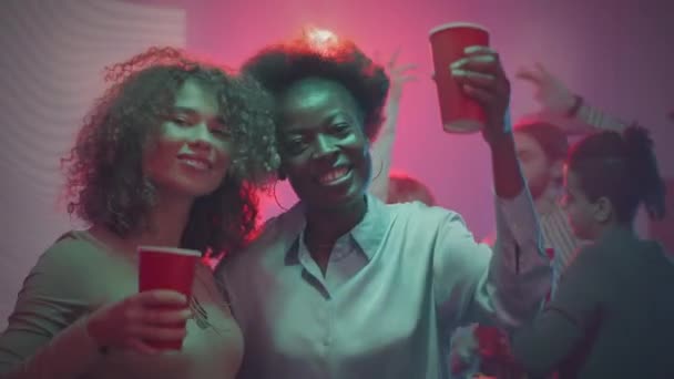 2人の若い魅力的な多民族女性の友人の中型肖像画は プラスチック製の赤いカップで乾杯し カメラで微笑み ナイトクラブでパーティーで友達と楽しんでいます — ストック動画