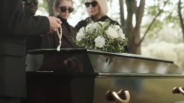 在公墓举行的户外丧礼上 一群悲伤的人们手持白念珠站在黑色棺材和牧师身旁 默念着祈祷 — 图库视频影像