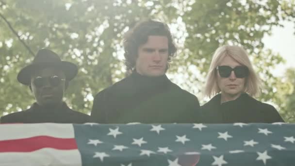 アウトドアの葬儀の際に悲しみ嘆き悲しみ嘆き悲しむアメリカの埋葬旗でケケットで立っている黒い服の多様な人々 — ストック動画