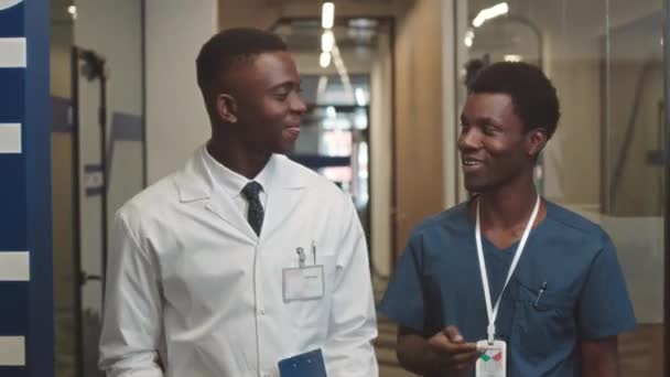 若いアフリカ系アメリカ人医師と彼のアシスタントが現代の診療所を歩きながら話しながら腰を追跡し 患者の検査結果を議論 — ストック動画