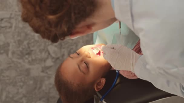 口を開いた歯科椅子に座っている子供の患者の歯を調べながら 女性歯科医の垂直ショット — ストック動画