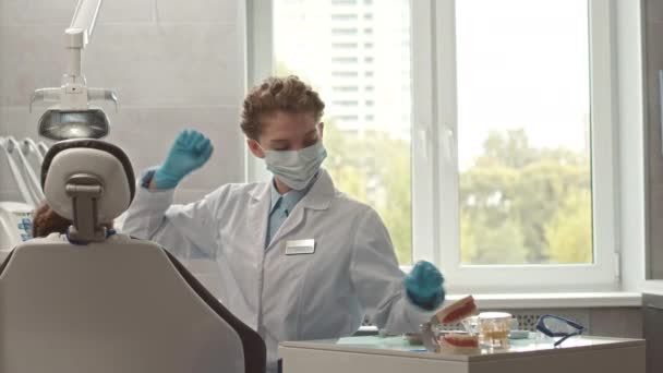 白い実験室のコート 使い捨て可能な表面のマスクおよび手袋の若い白人女性歯科医歯科椅子に座っている子供の患者の歯科検査をしている間 — ストック動画
