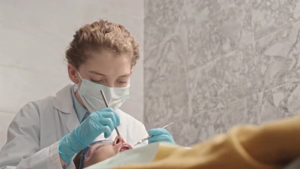 若い白人女性歯科医 顔面マスクと手袋 無菌手袋 クリニックで歯科治療中に患者歯を調べながら — ストック動画