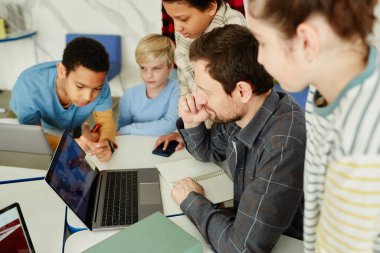 Okulda dizüstü bilgisayar kullanan bir grup çocukla erkek öğretmenin yüksek açılı portresi.