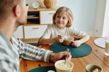 Baba ve oğlunun sabah birlikte kahvaltı ederken çekilmiş yüksek açılı portresi. Gülümseyen sarışın çocuğa odaklanmış.