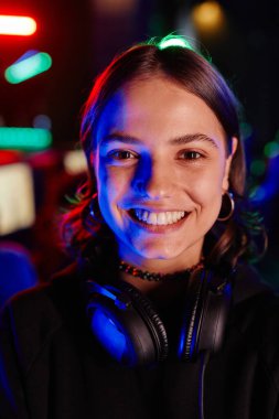 Siber spor takımındaki genç bir kadının dikey yakın plan portresi neon ışıklarıyla neşeyle kameraya gülümsüyor.