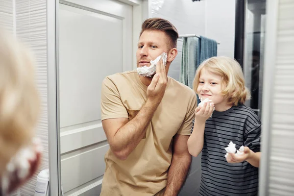 Portrait of father and son shaving together looking in mirror during morning routine in bathroom
