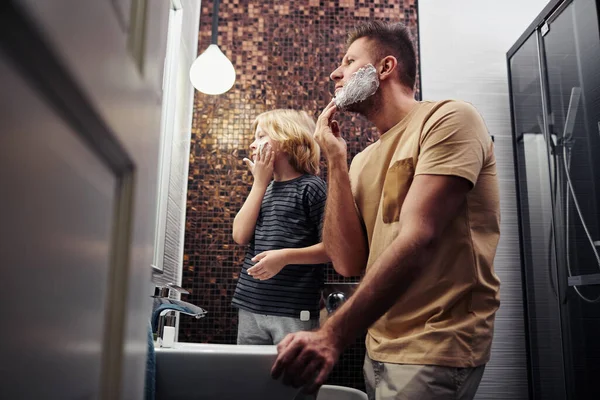 Side view portrait of father and son shaving together in bathroom and looking at mirror
