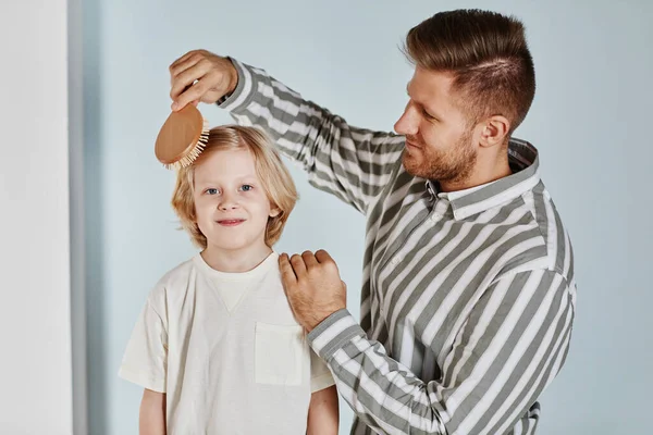 Waist up portrait of caring father brushing hair of cute young boy while getting dressed together
