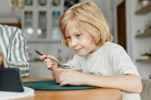 年轻金发男孩在餐桌边使用智能手机和在线观看视频的肖像 复制空间 — 图库照片