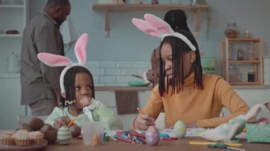 Tavşan kulaklığı takan iki Afrikalı Amerikalı kız kardeşin bel hizasında paskalya bayramını aileleriyle birlikte mutfak masasında el işi yaparak kutluyorlar.