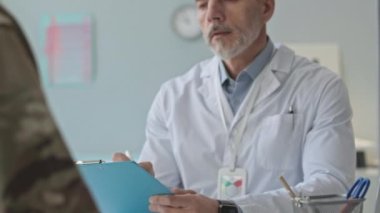 Kafkasyalı orta yaşlı askeri doktorun hastane randevusu sırasında askerin sağlığıyla ilgili konuşmalarını dinlerken tıbbi belgelere not alması.