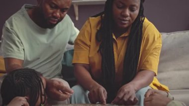 Afrikalı Amerikalı evli çift, modern oturma odasındaki kahve masasının üzerindeki yapbozu çözmek için iki küçük kızına yardım ediyor.
