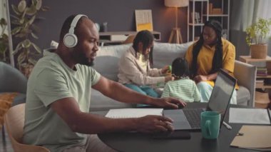 Eşi ve iki küçük kızı arka planda koltukta oyuncaklarla oynarken, ev bilgisayarında çalışan, kablosuz kulaklık takan Afrikalı Amerikalı bir adamın belden yukarısı görüntüsü.