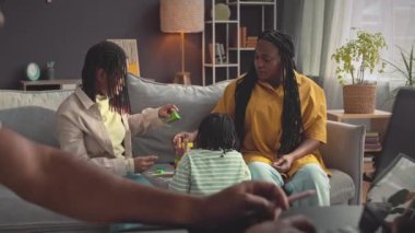 Siyah bir kadın, iki küçük kızıyla oturma odasında kanepede oynarken kocası da bilgisayarla evden çalışıyor.