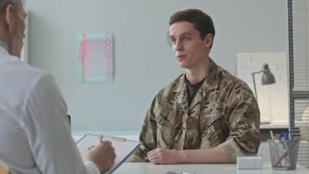 若い白人兵士が軍服を着て病院で診察任命中に医者と話すのを待つ — ストック動画
