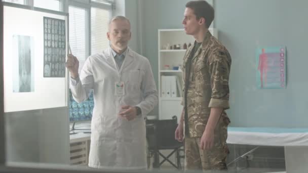 白いラボコートの白いラボコートの高齢者の医師の中型ショットは 医師の診察中に医師のオフィスに立っている若い兵士にMriスキャンを示しました — ストック動画