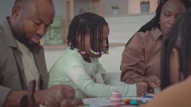 İki çocuklu modern siyahi aile mutfak masasında oturmuş Paskalya süslemeleri yapıyor, kartpostal çiziyor ve yumurta boyuyor.