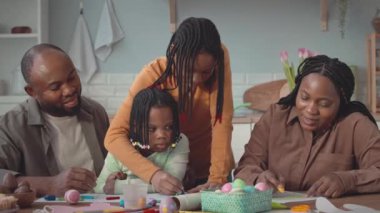 Dört kişilik mutlu siyah aile Paskalya süslemelerini modern mutfakta yapıyorlar, kartpostallar çiziyorlar ve paskalya yumurtalarını boyuyorlar.