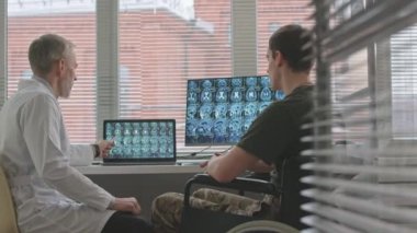 Orta boy, tekerlekli sandalyede oturan beyaz askerin beyin MR 'ı ekranda askeri doktorla tartışırken görüntüsü.