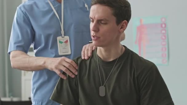 医師室の若い白人兵士の負傷した肩を調べる青いスクラブの認識できない理学療法士 — ストック動画