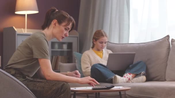 年轻的白人女军人数着家里的军费 而她十几岁的女儿则在舒适的客厅里用笔记本电脑在沙发上干活 — 图库视频影像