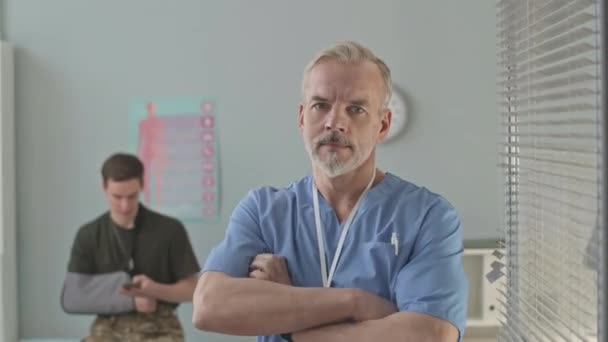 中年白种人军医的中肖像画 双手交叉在明亮的现代办公室里摆出拍照姿势 年轻士兵的手臂骨折坐在医疗沙发上作为背景 — 图库视频影像