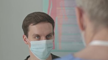 Klinikteki check-up randevusu sırasında yüz maskesi takmış genç beyaz adamın omuz üstünden doktorla konuşması.