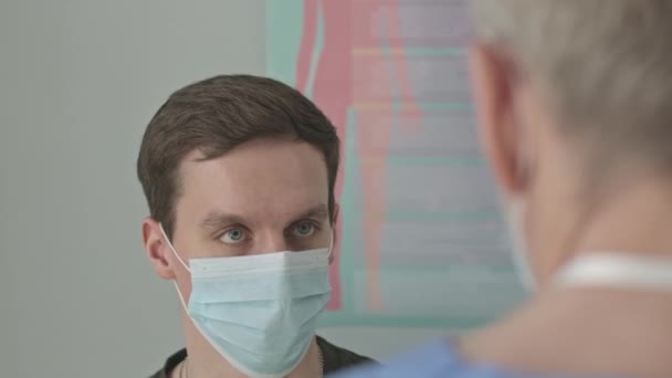 診療所の診察予約中に医者に話しかける顔のマスクの若い白人男性の肩のショットに — ストック動画