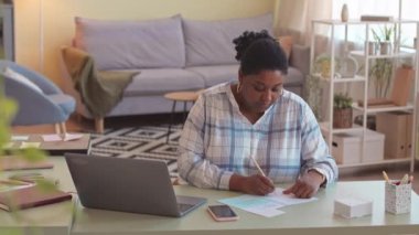 Genç ve cüsseli Afro-Amerikan kadın evde çalışıyor, modern oturma odasında dizüstü bilgisayarın önünde oturuyor ve not defterindeki notlara bakıyor.