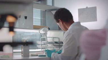 Beyaz laboratuvar önlüğü ve eldivenleri giyen odaklanmış genç, çift ırklı erkek bilim adamının beli Petri kabında tanınmayan sıvı maddeyi analiz ederken iş arkadaşlarıyla birlikte araştırma laboratuarında çalışıyor.