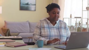 Genç, kıvrımlı, Afro-Amerikan iş kadını oturma odasındaki masada dizüstü bilgisayarda çalışıyor.