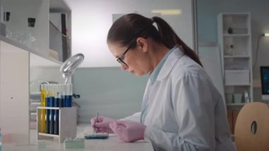 Beyaz laboratuvar önlüğü ve gözlük takmış, petri kabında kültürü incelerken cımbız ve cam parçası kullanan konsantre olmuş beyaz kadın mikrobiyoloğun belden yukarı görüntüsü.