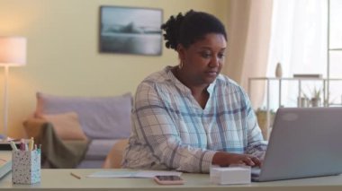 Genç, kıvrımlı, siyah bir kadın rahat oturma odasında oturmuş dizüstü bilgisayarla sohbet ediyor.