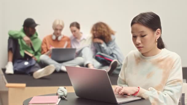 在大学时 年轻漂亮的亚洲女学生坐在笔记本电脑上学习 而她的同学则坐在地板上 在模糊的背景下聊天 学习速度缓慢 — 图库视频影像