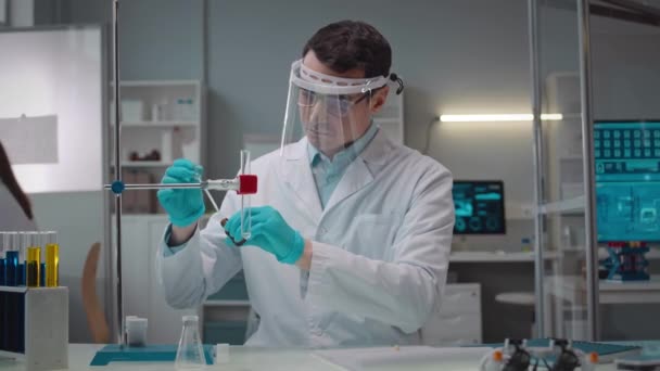 年轻白种人男性科学家身穿白衣 戴着防护面罩在现代实验室进行实验 在玻璃管中插入一些无法辨认的红色液体的起跳镜头 — 图库视频影像